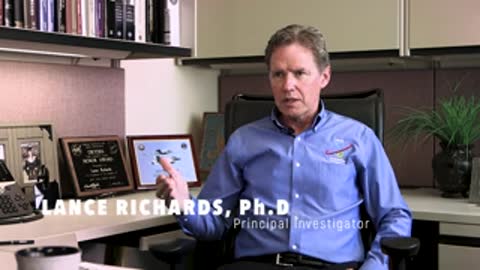 A man sitting in an office speaking, with computer monitors and shelves behind him.