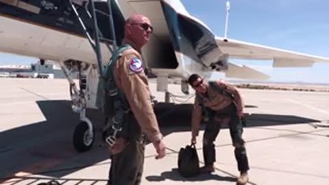 Two pilots in flight gear standing outside near a parked aircraft.