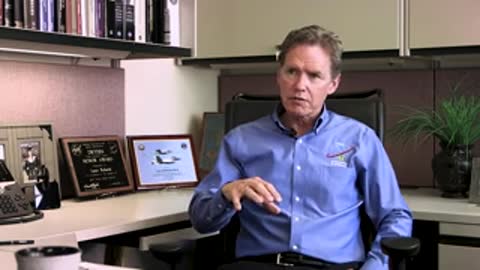 A man in a blue shirt gesturing while speaking in an office interview.