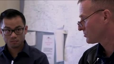 Two men wearing glasses looking down and discussing documents indoors.