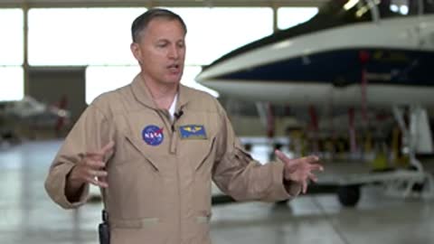 A man in a flight suit talking and gesturing inside an aircraft hangar.