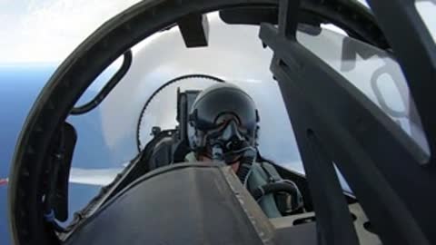 A pilot sitting inside a fighter jet cockpit under a bright sky.