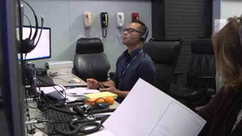 Two people working at desks with computers in a mission control environment.