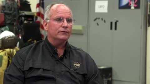 Man in black shirt talking indoors in front of a white wall.