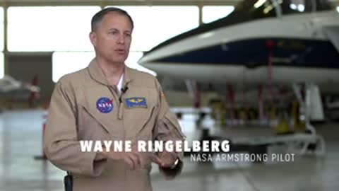 A man wearing a flight suit speaking, with an aircraft in the background.