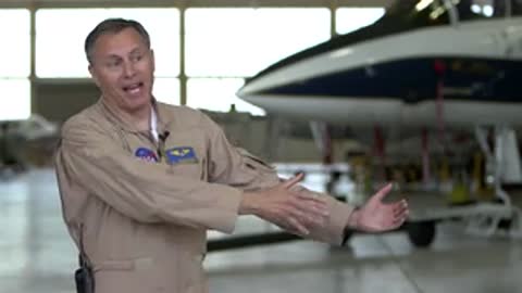 Pilot in flight suit gesturing while speaking in an aircraft hangar.