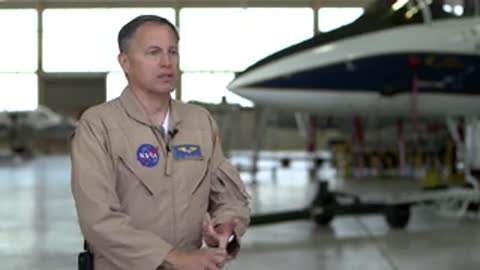 Man in flight suit speaking in front of an aircraft in a hangar.
