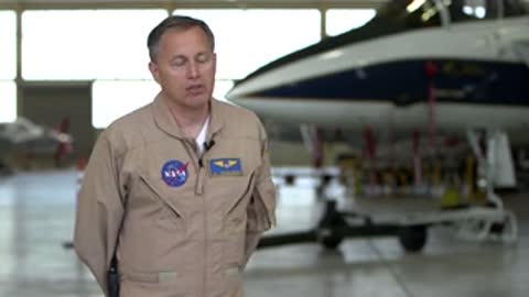 A man in a flight suit talking in front of an airplane hangar.