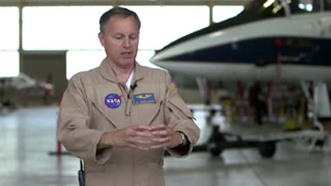 Man in beige jumpsuit talking in front of aircraft.