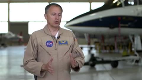 Man in beige jumpsuit talking in hangar.