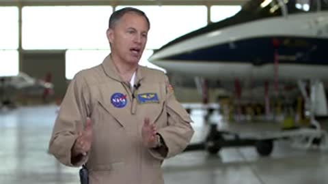 Man in beige jumpsuit speaking in hangar.