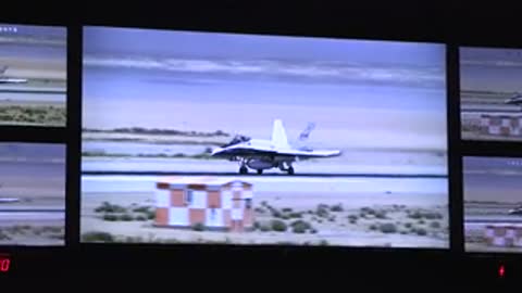 Multiple monitors displaying footage of a fighter jet on a runway.