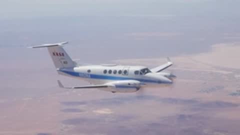 A twin-propeller aircraft flying in the sky over a dry desert landscape.