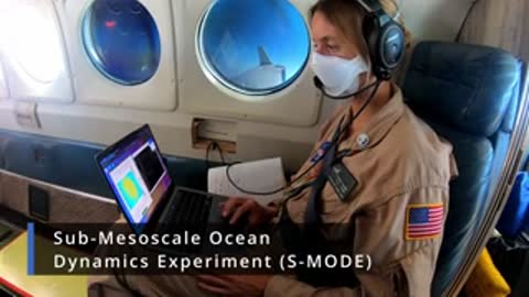 A person wearing a face mask and headphones sitting inside a plane using a laptop.