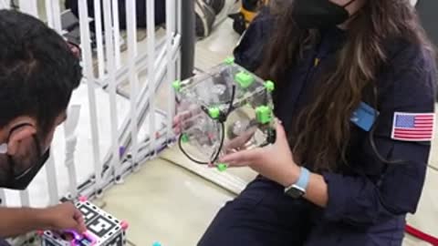 A person holding a small cube-shaped satellite structure inside a testing facility.