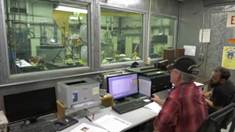 Control room staff monitoring computer displays through large observation windows.