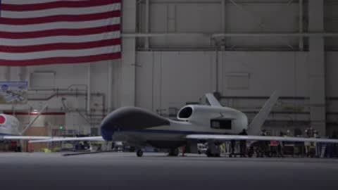 A large drone parked inside a hangar with an American flag behind.