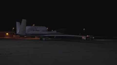 A large drone parked on a tarmac at night under exterior lighting.