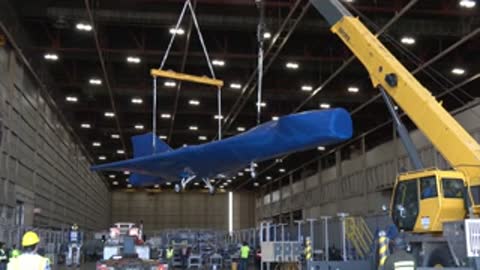 Covered aircraft being lifted by a yellow crane inside a large industrial facility
