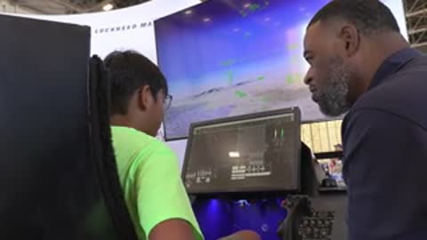 A man talks to a boy playing a flight simulator inside a large room.