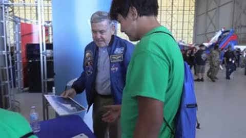 An astronaut in uniform signs autographs for people at a table inside.