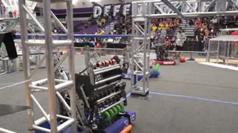 A small robot moves around a netted arena while people watch in the background.