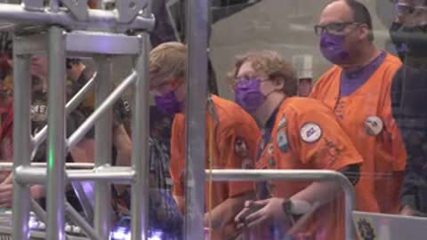 People wearing orange vests watch a robotics competition through clear protective barriers.