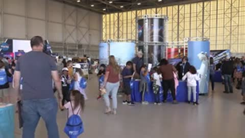 People walk across a large exhibition hall floor with booths and displays.