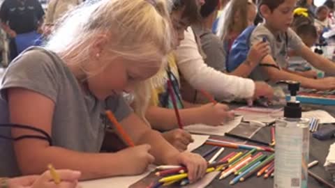 Children sit at tables colouring with markers in a large indoor event space.