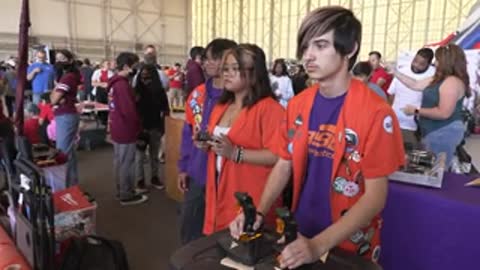 Two teenagers play a video game using controllers while standing in a crowd.