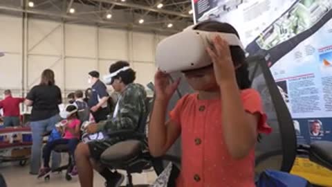 Children put on and adjust VR headsets while sitting in chairs at an event.