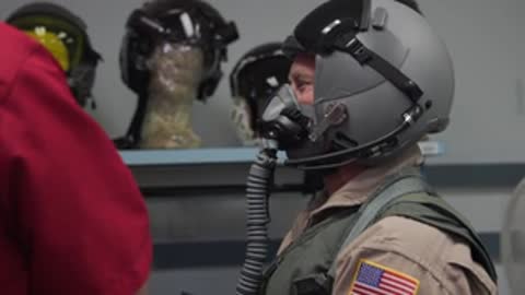 Pilot in flight gear and helmet standing in front of equipment racks.
