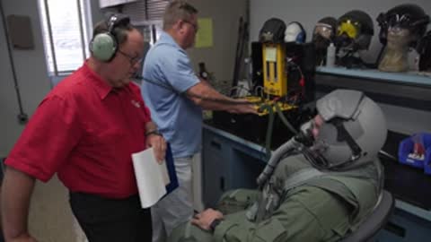 Researchers in a lab reviewing equipment with a suited subject seated nearby.
