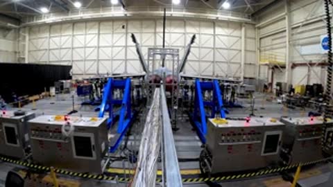View from behind an aircraft structure being tested within a large indoor laboratory setting.