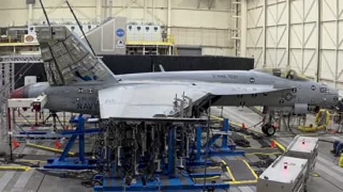 Side profile of an aircraft mounted on hydraulic test rigs inside a brightly lit hangar.