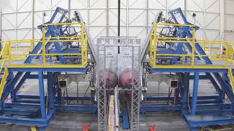 Rear view of an aircraft with its control surfaces elevated inside a testing facility hangar.