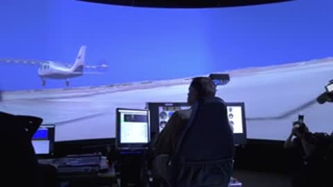 Flight simulator control station with personnel monitoring screens displaying aircraft flight simulations.