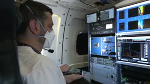 A crew member wearing a mask sits at a workstation inside an aircraft.