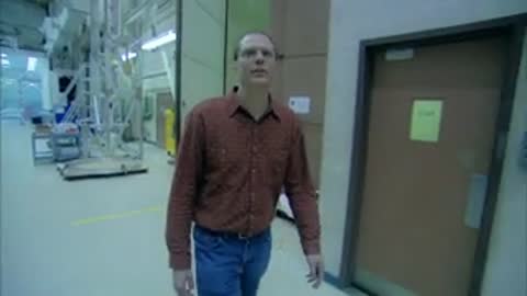 Man walking down a cleanroom hallway, identified by on-screen text as Kevin Boyce