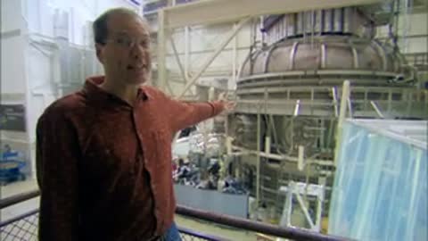 Man standing on a balcony gesturing toward a large vacuum chamber