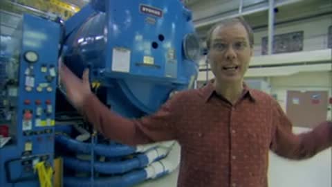 A medium shot of a man speaking in front of a blue pressure vessel chamber.