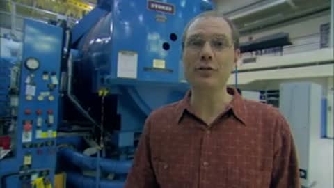 A medium shot of a man speaking inside the facility near a blue vacuum chamber.