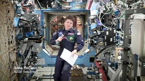 An astronaut holding a microphone and papers, speaking