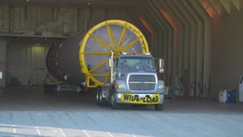 Medium tracking shot of a semi-truck carrying a rocket stage exiting a building.
