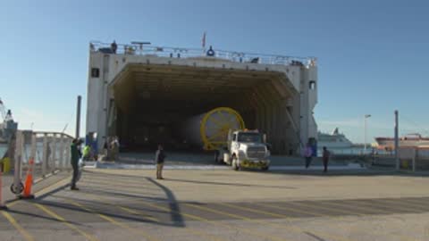 Wide shot of a semi-truck backing a rocket stage out of a large cargo vessel.