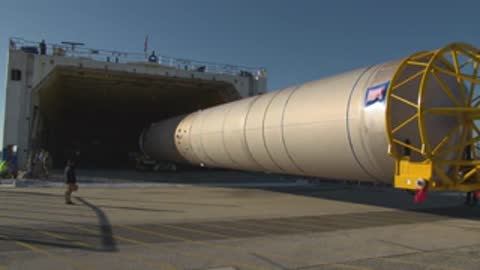 Medium tracking shot of a long rocket stage being towed out of a cargo vessel.