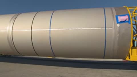 Wide tracking shot of a long rocket stage being transported on a truck outdoors.