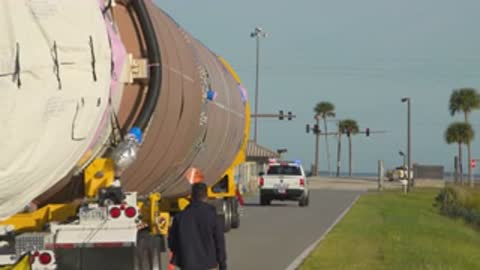 Medium tracking shot of a large rocket stage being transported down a road.