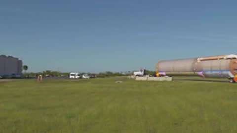 A wide shot shows a truck transporting a rocket booster across a grassy field.