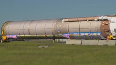 A wide shot shows a long rocket booster being transported by a truck on a road.